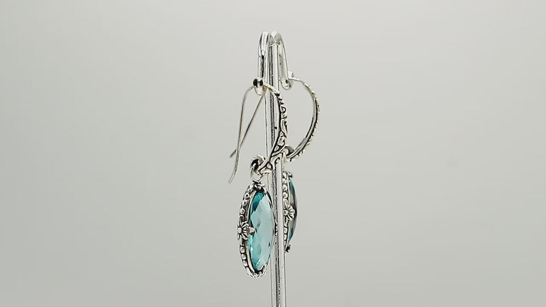 A handcrafted pair of 925 sterling silver earrings featuring faceted oval Blue Topaz Quartz stones with detailed Balinese floral motifs and secure earwire hook design.